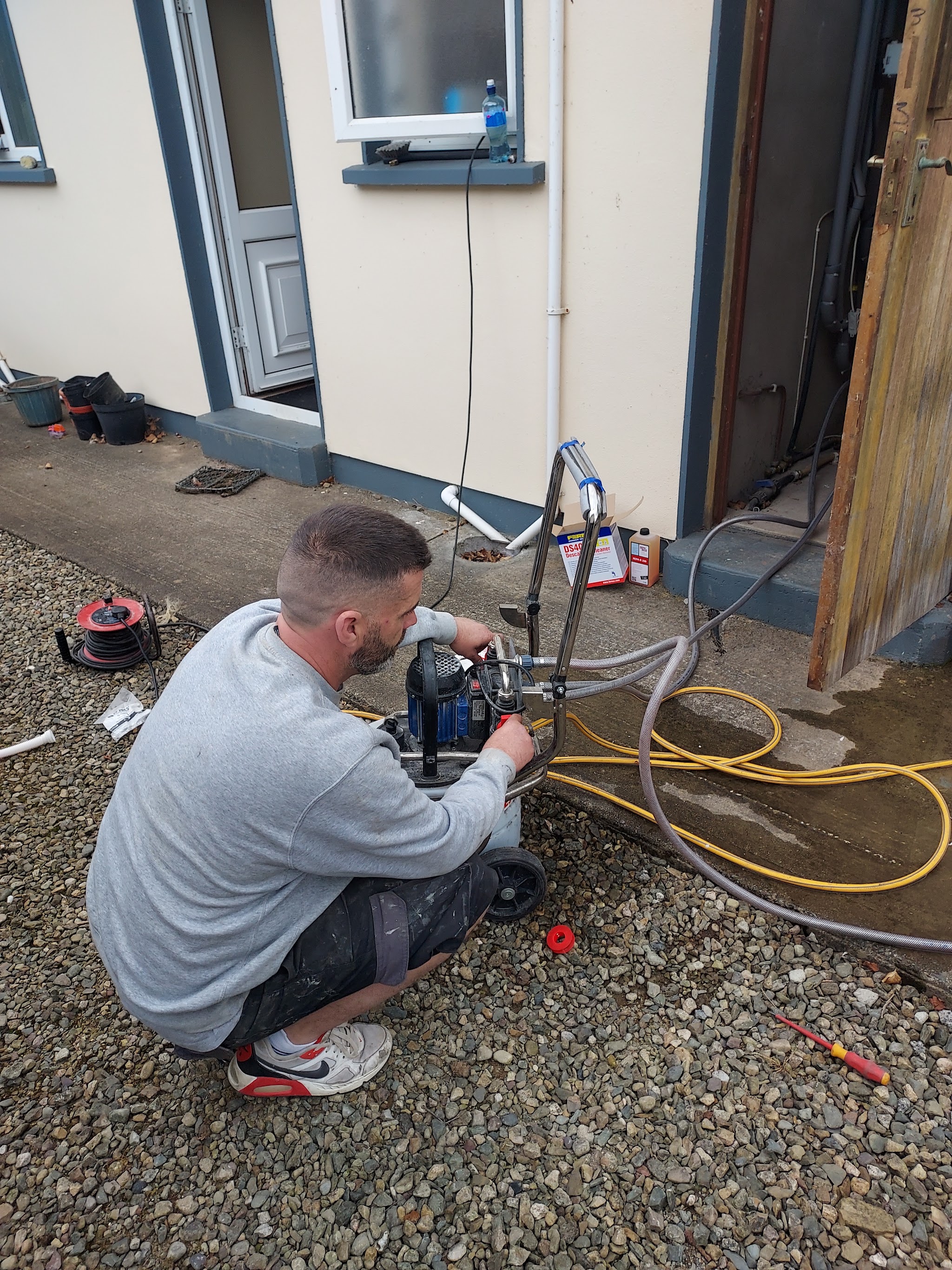 Plumber kneeling outside a house, working a machine connected to hoses on the gravel